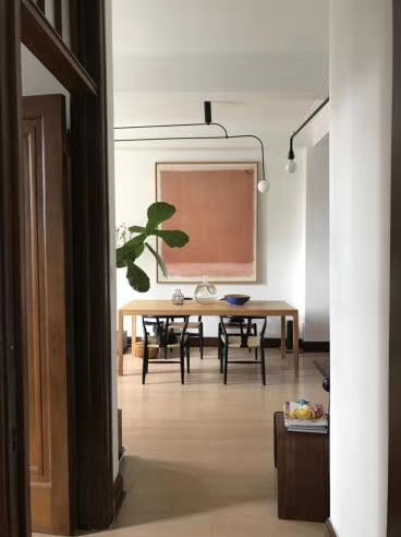View into a bright dining room: a wooden table with chairs, a large leafy plant, and a pink abstract painting on the wall above.