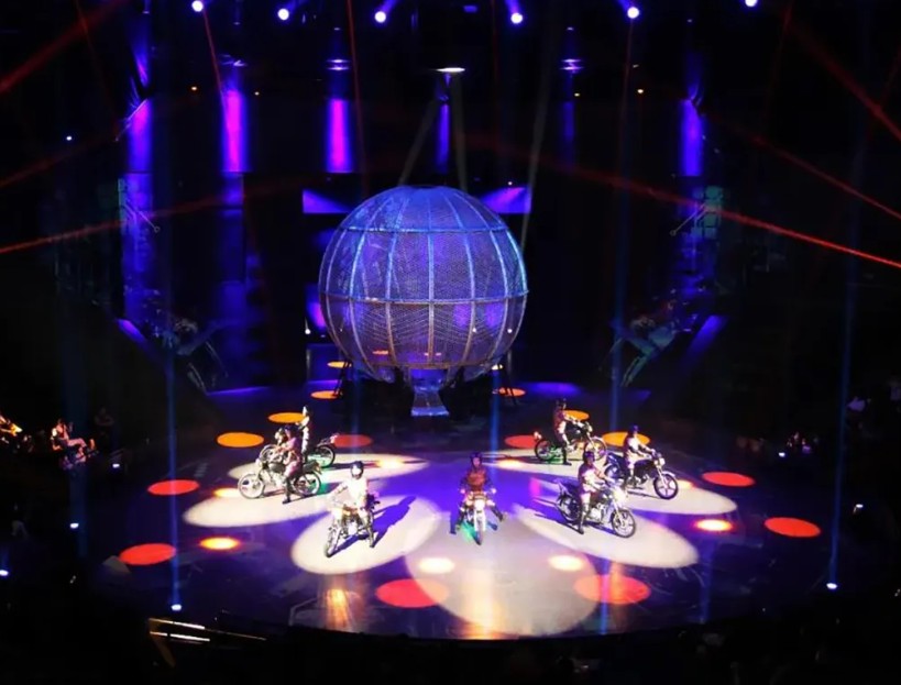 Multiple motorcycles race simultaneously inside a steel sphere only six meters in diameter. The riders cross paths with centimeters of clearance, engines roaring, lights flashing.